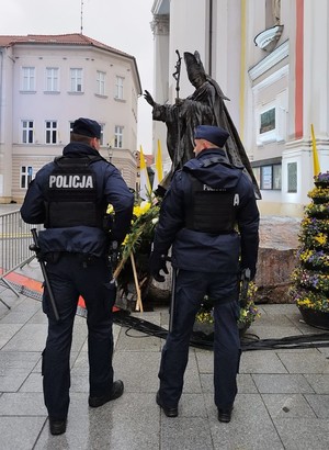 Dwóch umundurowanych policjantów stojących tyłem. W tle pomnik Jana Pawła II i kościół.