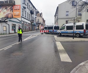 Umundurowany policjant ruchu drogowego stojący na skrzyżowaniu. W tle oznakowane radiowozy i budynki.