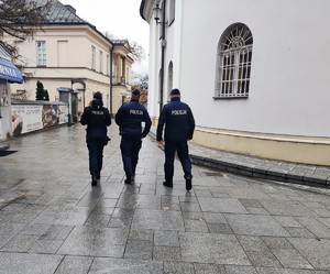 Umundurowana policjantka i dwóch umundurowanych policjantów idących między budynkami. osoby są tyłem.