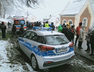 Na zdjęciu widać oznakowany radiowóz orz karetkę pogotowia. W tle tłum osób oraz umundurowany policjant i budynki.