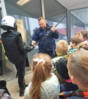 Na zdjęciu widać umundurowanego policjanta, który trzyma tonfę policyjną, obok stoi osoba, która ma załojony kask policyjny oraz kamizelkę kuloodporną. Wokoło grupa dzieci.