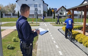 Na zdjęciu widać dwóch umundurowanych policjantów, którzy oceniają jazdę dwóch osób na rowerze po specjalnie przygotowanym torze ze znakami drogowymi. W tle domy i samochód.