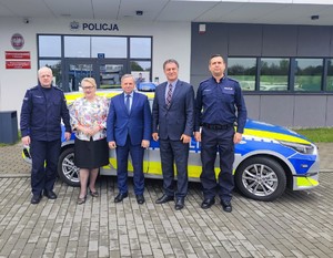 Na tle budynku wadowickiej komendy od lewej strony stoi umundurowany policjant, kobieta i dwóch mężczyzn w stroju cywilnym oraz umundurowany funkcjonariusz Policji. Za nimi nowy oznakowany radiowóz kia ceed.