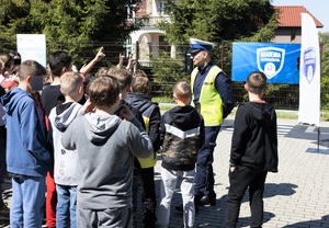 Umundurowany policjant ruchu drogowego w kamizelce odblaskowej z napisem Policja. Przed nim stoi grupa dzieci, część z nich podnosi rękę do góry.