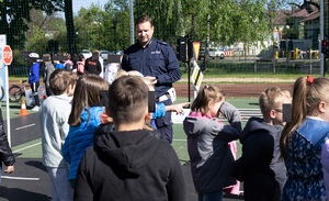 Umundurowany policjant stojący przodem, przed nim grupa dzieci. W tle również widać dzieci oraz znaki drogowe. Osoby znajdują się na boisku szkolnym.