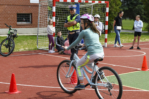 Na zdjęciu dziewczynka jadąca na rowerze po wyznaczonym torze. Obok umundurowany policjant, w tle grupa dzieci. Osoby znajdują się na boisku sportowym.