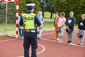 Na zdjęciu umundurowany policjant w kamizelce odblaskowej z napisem Policja, stojący tyłem. Przed nimi grupa dzieci. Osoby znajdują się na boisku sportowym.