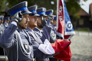 Czterech umundurowanych policjantów stojących obok siebie oddających honor.  Jeden z Funkcjonariuszy trzyma złożona flagę Polski, drugi sztandar.