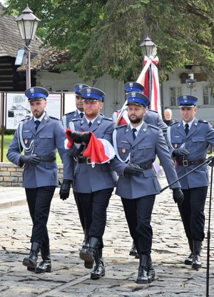 Na zdjęciu widać sześciu umundurowanych policjantów idących w dwuszeregu. Z tyłu policjant idący w środku trzyma sztandar, z przodu policjant idący w środku niesie złożoną flagę Polski.