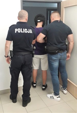 Na zdjęciu po lewej stronie stoi umundurowany policjant, po prawej stronie policjant w stroju cywilnym, którzy trzymają zatrzymanego mężczyznę, który ma założone kajdanki zespolone i kask ochronny na głowę. Osoby są tyłem, stoją w drzwiach.