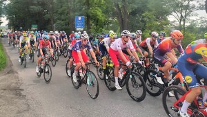 Na zdjęciu widać grupę rowerzystów uczestniczących w wyścigu kolarskim. Za nimi jadą samochody.