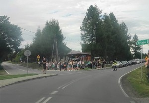 Umundurowany policjant ruchu drogowego stojący na skrzyżowaniu, kieruje ruchem. W tle stojące samochody, grupa ludzi i zabudowania.