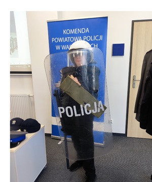 Chłopiec ubrany w kamizelkę kuloodporną i kask, w ręku trzyma pałkę orz tarczę policyjną. Z nim baner policyjny z napisem Komenda Powiatowa Policji w Wadowicach.