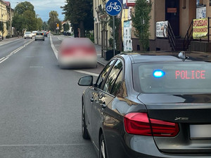 Na zdjęciu widać tył nieoznakowanego pojazdu policyjnego z podświetlonym na tylnej szybie napisem Policja. Przed nim kontrolowany na ulicy pojazd, samochód ten jest rozmyty na zdjęciu. Wokoło zabudowania.