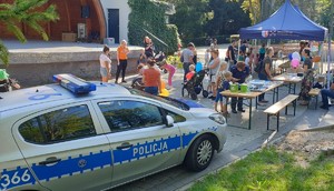 Na zdjęciu widać oznakowany radiowóz, po prawej stronie rozłożone namioty i stoliki na których są miedzy innymi ulotki i policyjny sprzęt. Wokoło grupa osób i umundurowana policjantka.