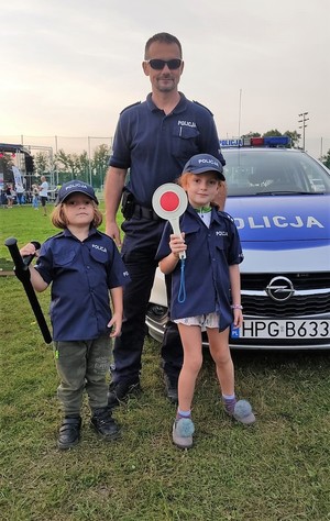 Umundurowany policjant, po prawej i po lewej stronie stoją małe dziewczynki ubrane w policyjny strój. Jedna trzyma tarcze sygnalizacyjną, druga tonfe. W tle policyjny radiowóz.