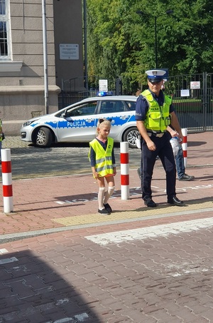 Mała dziewczynka w kamizelce odblaskowej przechodząca przez przejście dla pieszych z umundurowanym policjantem ruchu drogowego. W tle budynek i oznakowany radiowóz.