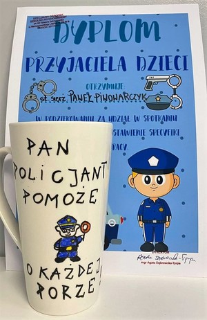 Na zdjęciu widać dyplom dla policjanta za udział w spotkaniu. Przed dyplomem jest postawiony kubek z napisem Pan policjant pomoże o każdej porze. Pośrodku jest narysowany policjant.