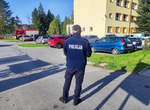 Na drodze wjazdowej do szkoły stoi tyłem umundurowany policjant. W tle widać budynek szkoły i zaparkowane samochody oraz wóz strażacki.