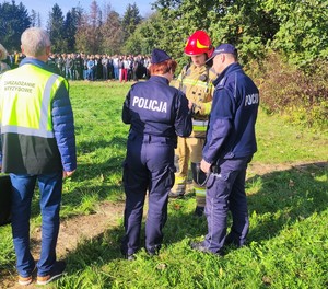 Na zdj zdjęciu widać umundurowaną policjantkę i policjanta, którzy stoją obok strażaka. Po lewej stronie stoi mężczyzna w odblaskowej kamizelce z napisem Zarządzanie kryzysowe. W tle stoi grupa osób. Wokoło drzewa i krzewy.