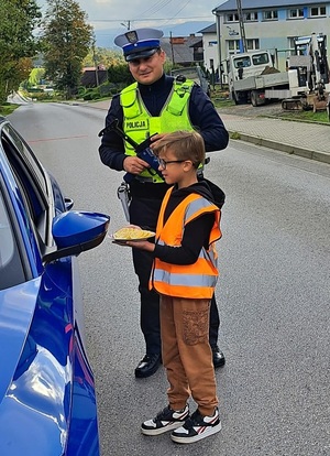 Umundurowany policjant ruchu drogowego, obok mały chłopiec w odblaskowej kamizelce który na talerzu ma plastry cytryny. Stoją na drodze obok niebieskiego samochodu.