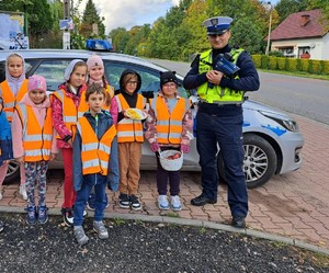 Grupa dzieci w kamizelkach odblaskowych stojących przodem na tle radiowozu. Po prawej stronie umundurowany policjnat  ruchu drogowego z miernikiem prędkości w ręce. W tle droga i budynek.