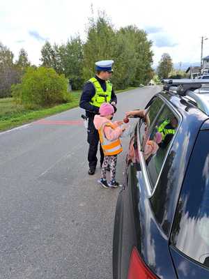 Umundurowany policjant ruchu drogowego i mała dziewczynka w kamizelce odblaskowej ,która ma koszyk z jabłkami. Po prawej stronie samochód, którego kierujący przez szybę zabiera z koszyka jabłko.