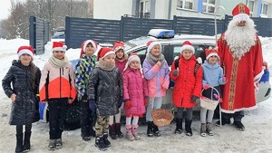 Na tle budynku szkoły i oznakowanego radiowozu stoi grupa dzieci w czapkach Mikołaja. Po prawej stronie stoi osoba przebrana za Świętego Mikołaja.