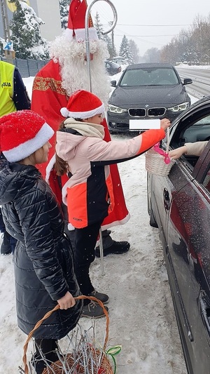 Na zdjęciu widać dwie dziewczynki w czapkach Mikołaja, które maja koszyczki. Obok stoi Mikołaj. Po prawej stronie fragment samochodu, którego kierowca zabiera z koszyka cukierek. W tle czarny samochód osobowy.