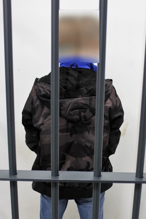 Zatrzymany mężczyzna  stojący tyłem w celi. Ubrany w kurtkę koloru ciemnego moro i niebieskie dżinsy.