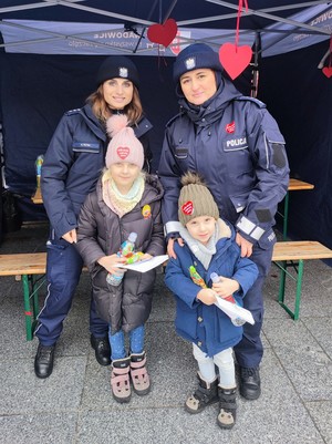 Dwie umundurowane policjantki. Przed nimi stoi dziewczynka i chłopiec. Osoby stoją na tle namiotu.