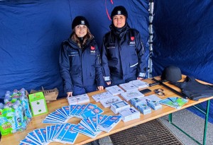 Dwie umundurowane policjantki. Przed nimi stolik na którym są poukładane różne ulotki edukacyjno informacyjne.