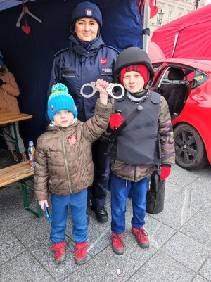 Umundurowana policjantka. Przed nią stoją dwaj chłopcy. Jeden trzyma kajdanki, drugi ma założone na sobie elementy umundurowania policyjnego.